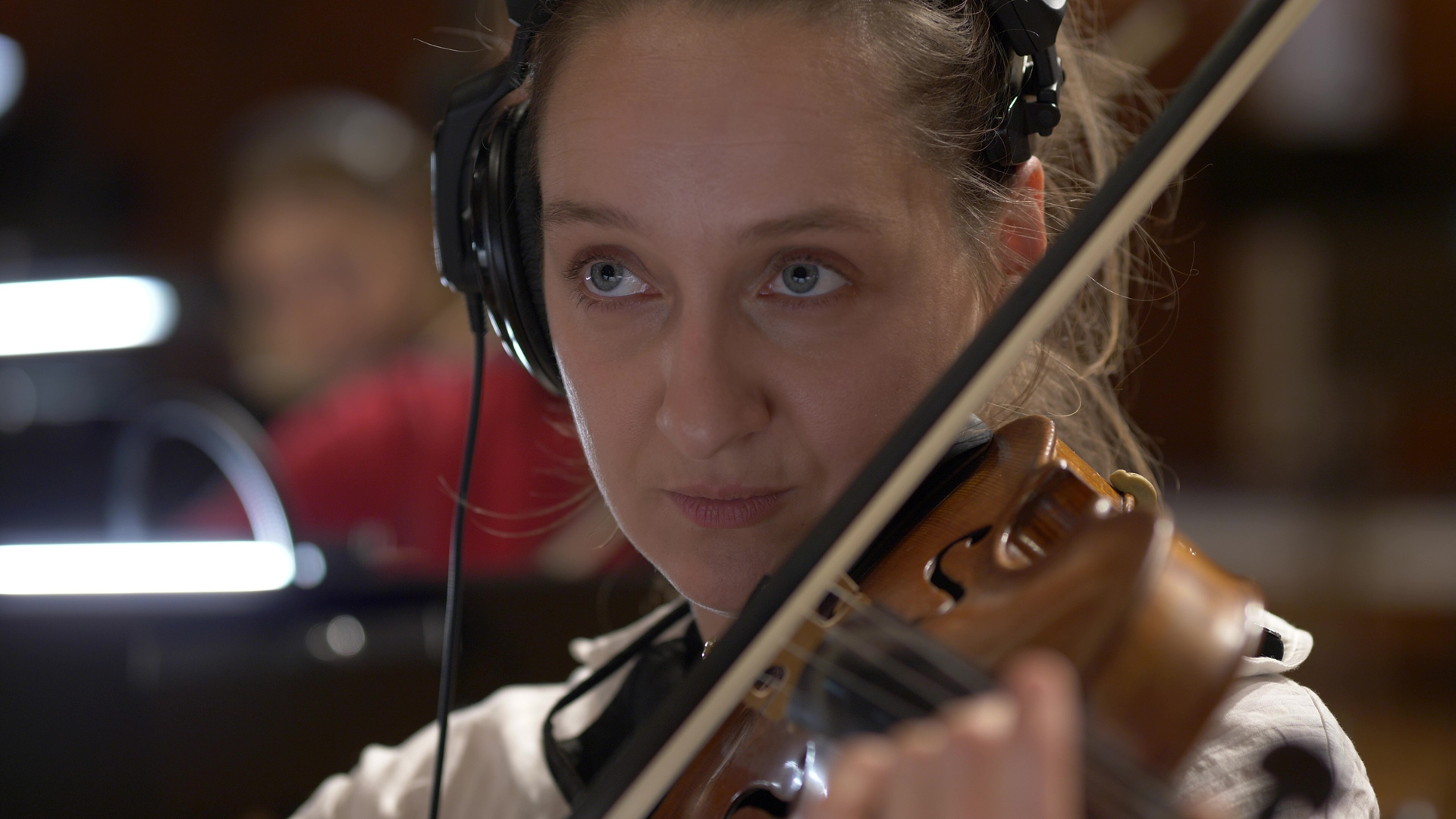 Close-up of a female violinist with light brown hair and blue eyes, wearing over-ear headphones, playing a violin during a recording session or orchestral performance.