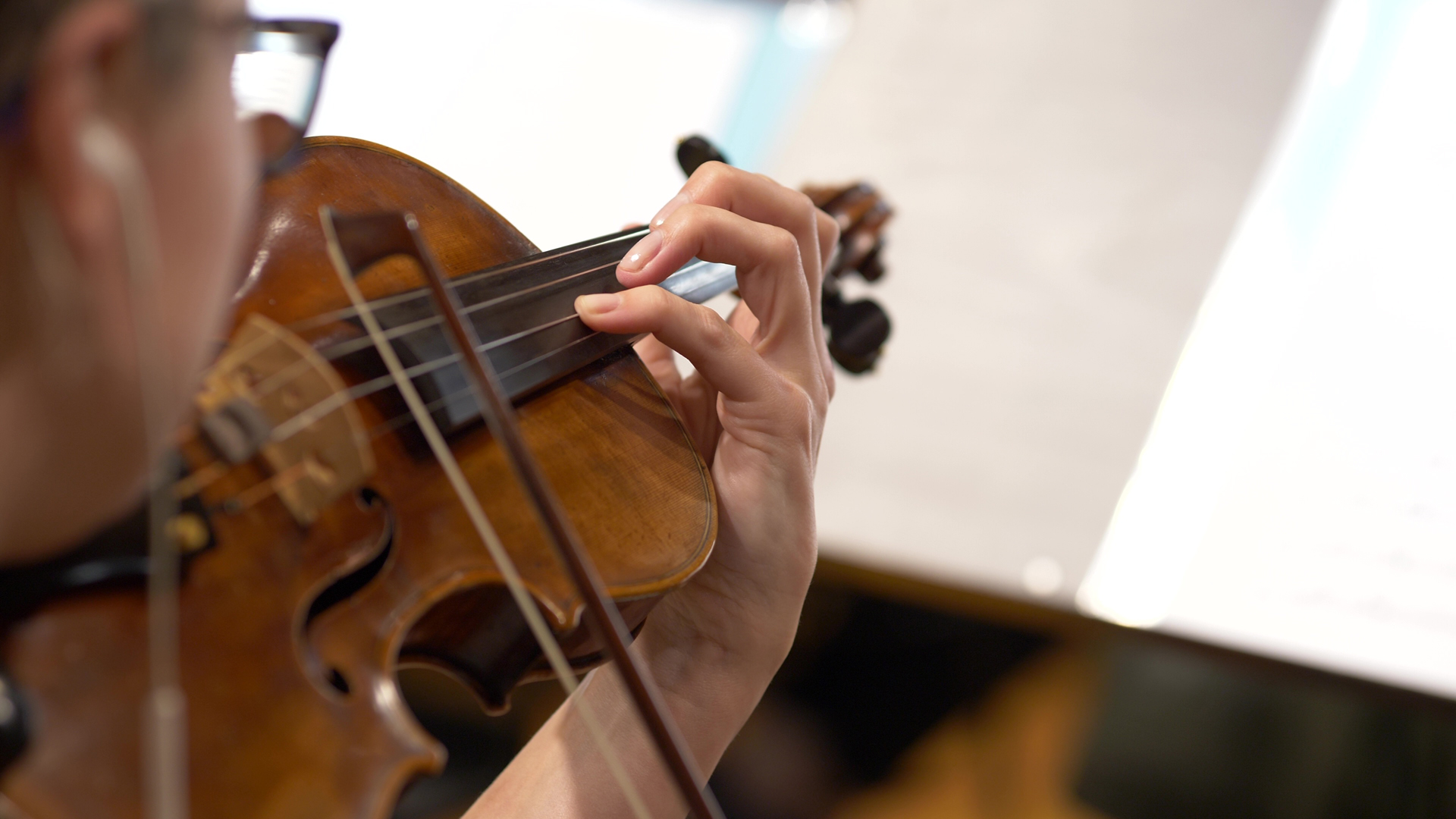 Close-up of a person wearing glasses playing a wooden violin with a bow during an orchestra performance, focusing on their hand pressing the strings and the instrument's details like the f-holes, in a concert or practice setting.