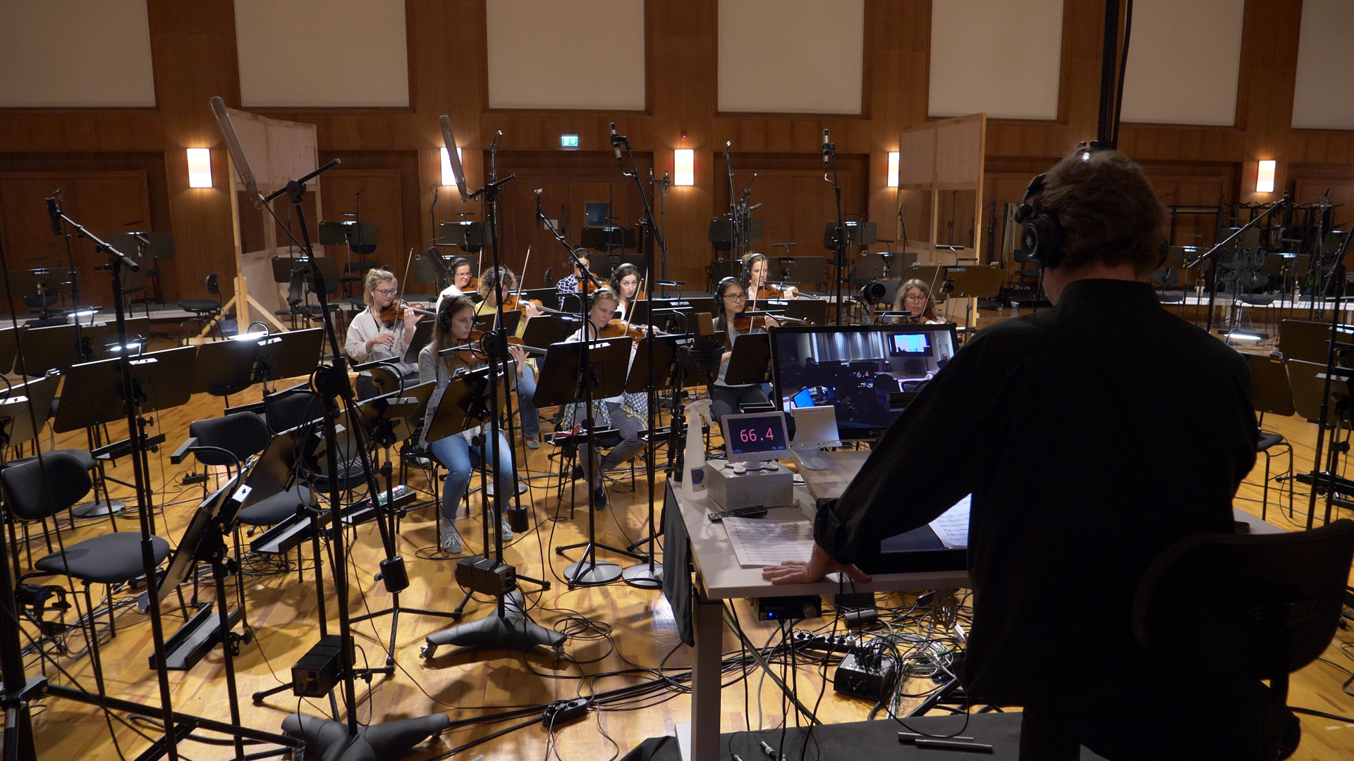 Orchestra recording session with musicians playing violins, microphones, and a sound engineer monitoring at a mixing desk.