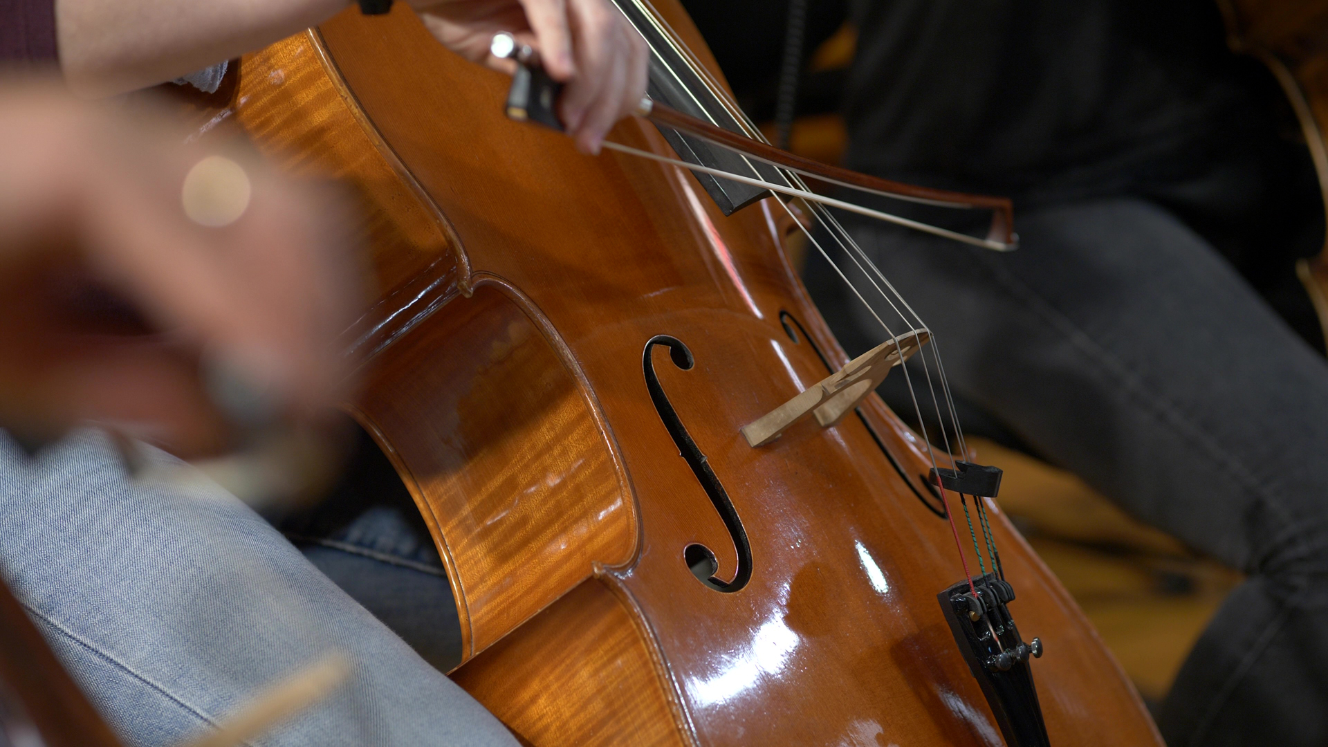 Close-up of a cello being played, showing the warm brown wood, f-holes, bridge, and strings. A musician's hands, wearing light blue jeans, are visible near the instrument, with a bow drawn across the strings by another player in the background.
