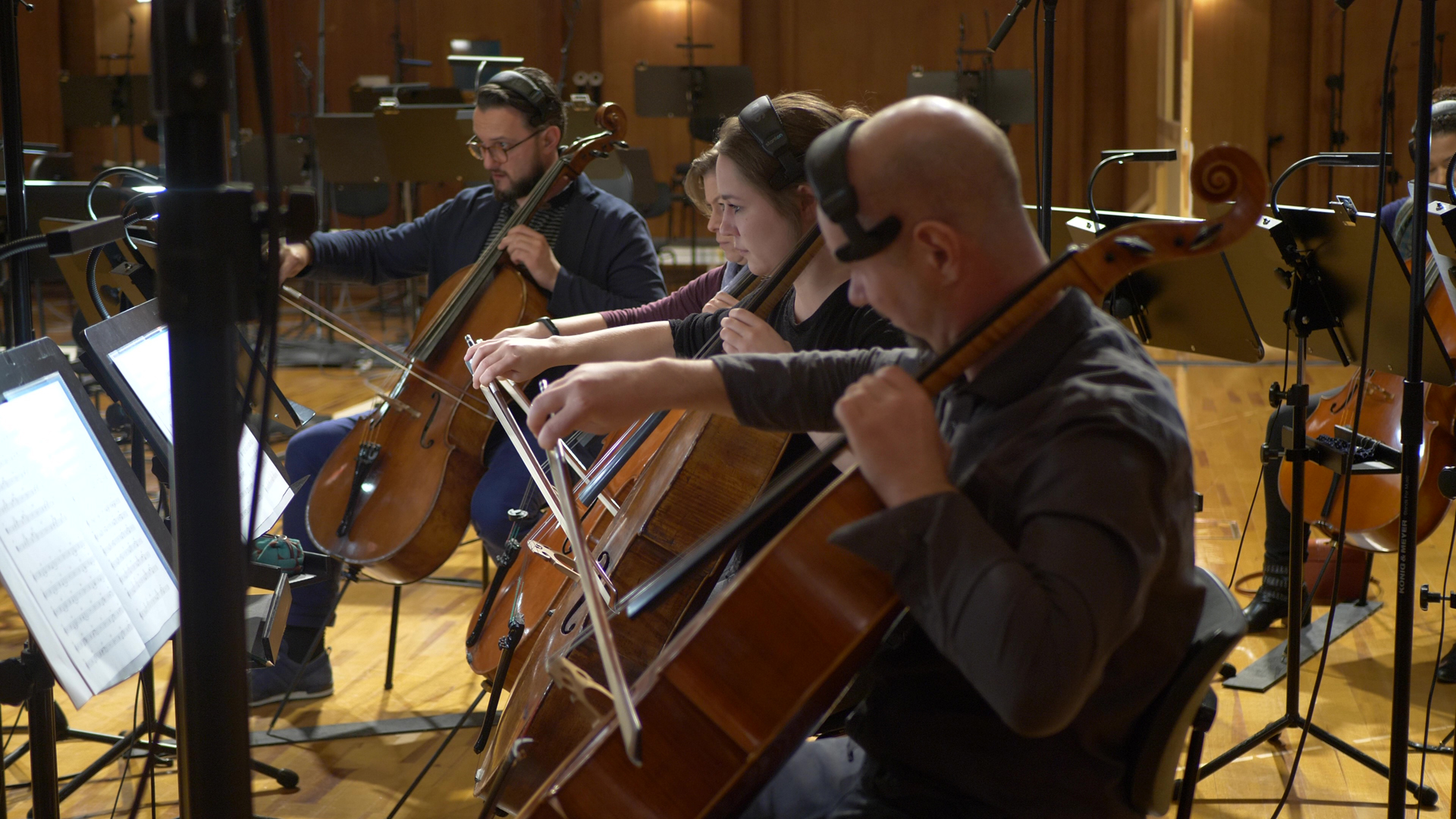 A group of four cellists, including a man with a beard and glasses, a woman, and another man in the foreground, playing cellos in a recording studio with sheet music stands and microphones.