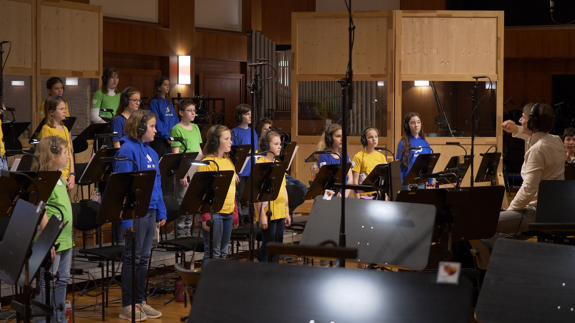 Choir of diverse children in yellow, blue, and green shirts with headphones, singing or recording in a studio with music stands and a conductor.