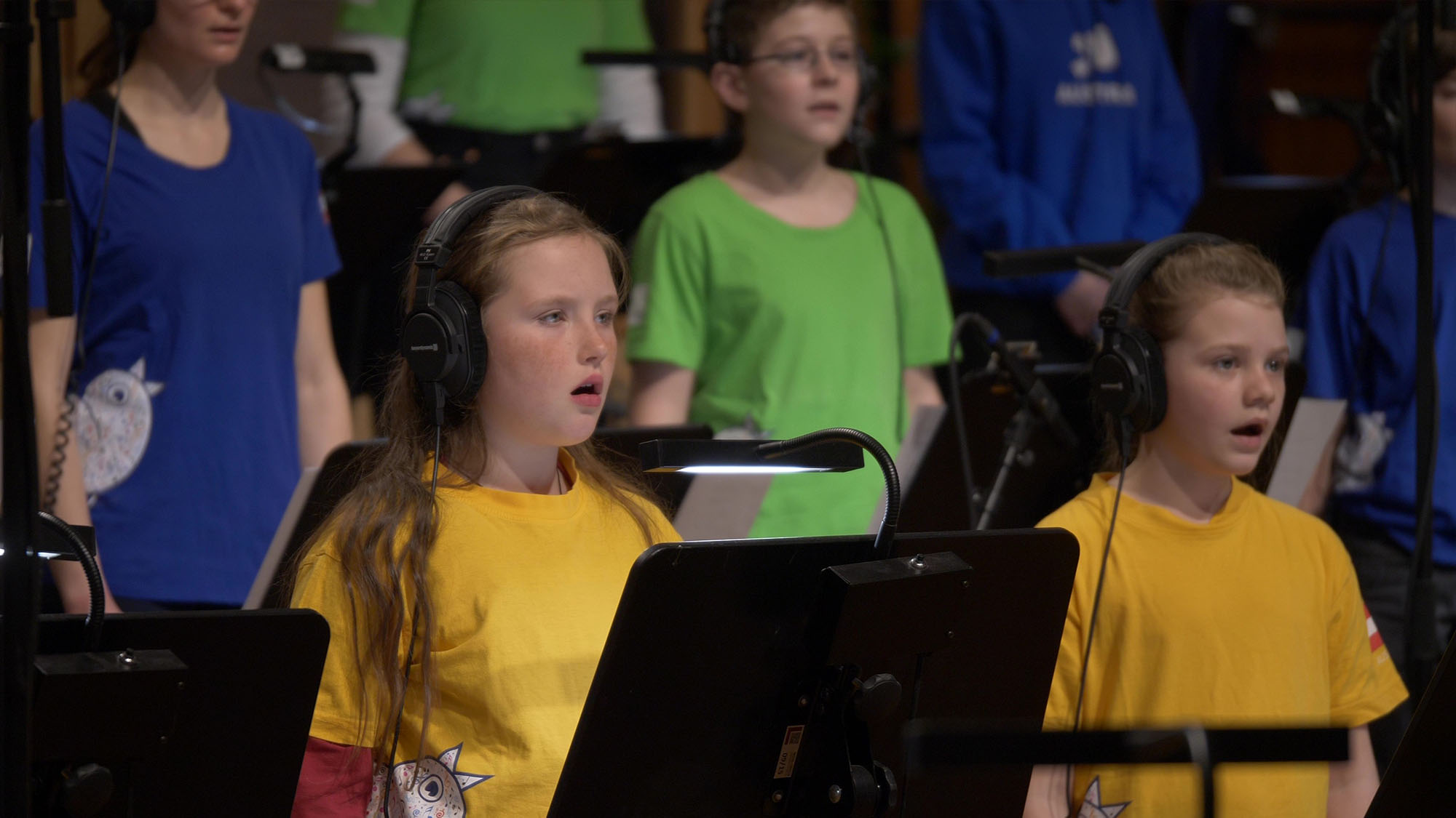 Headphones-wearing children singing in a choir with sheet music stands and lights, performing in a group.