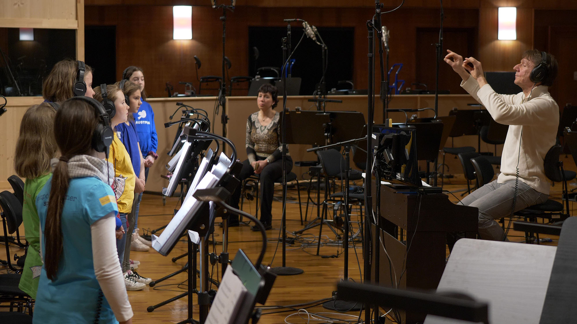 Music conductor with headphones directing a children's choir in a studio with microphones and music stands.