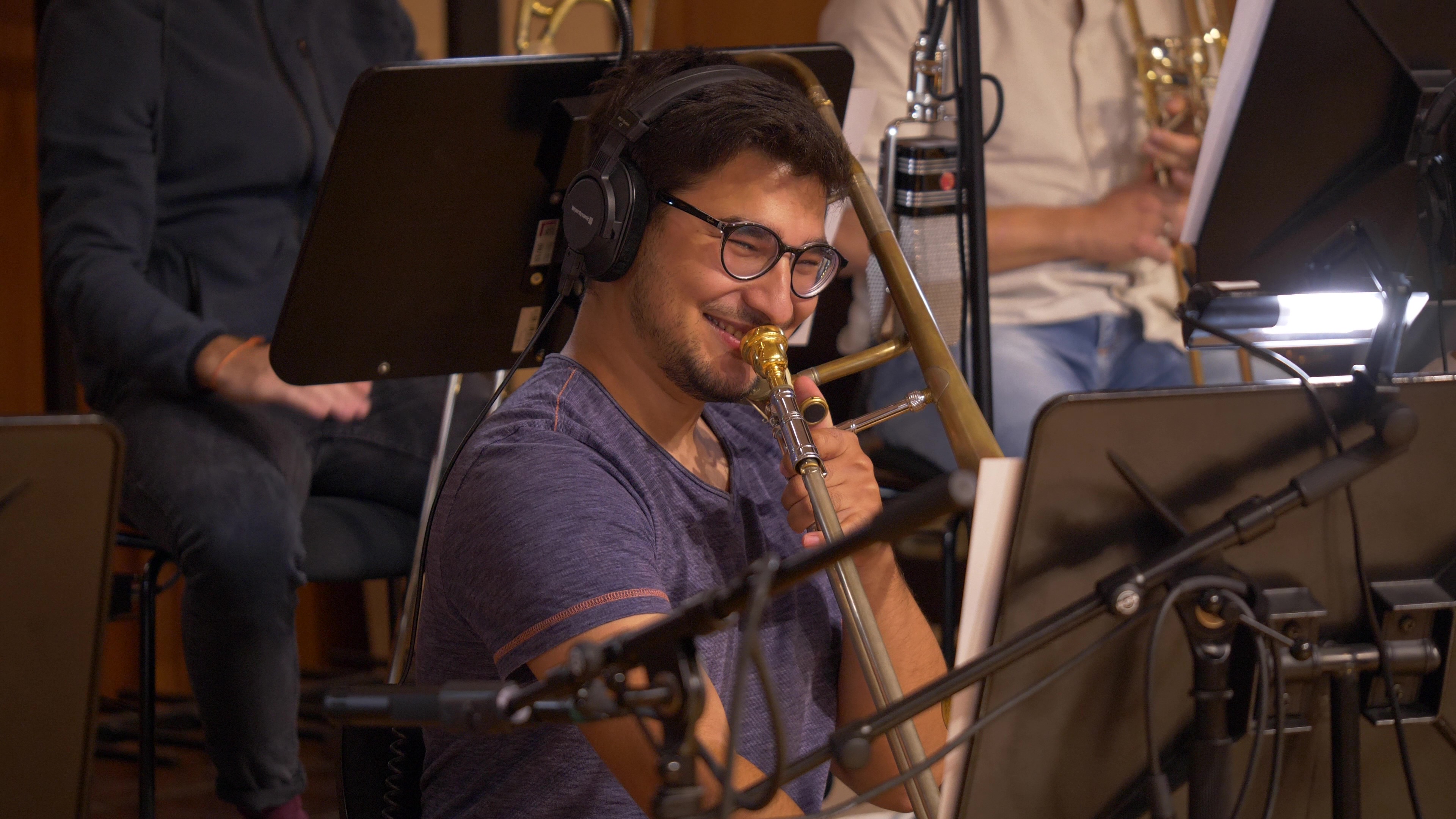 Smiling musician wearing headphones and glasses, playing a trombone, surrounded by a music stand and microphones in a recording studio.