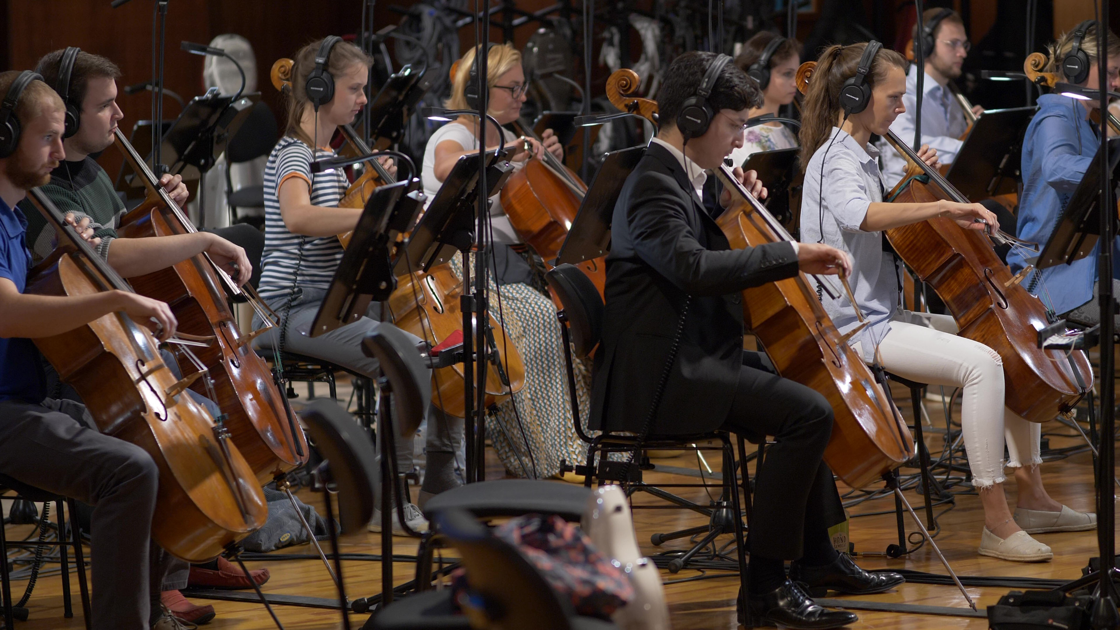 Musicians wearing headphones play cellos in an orchestra rehearsal or recording session.