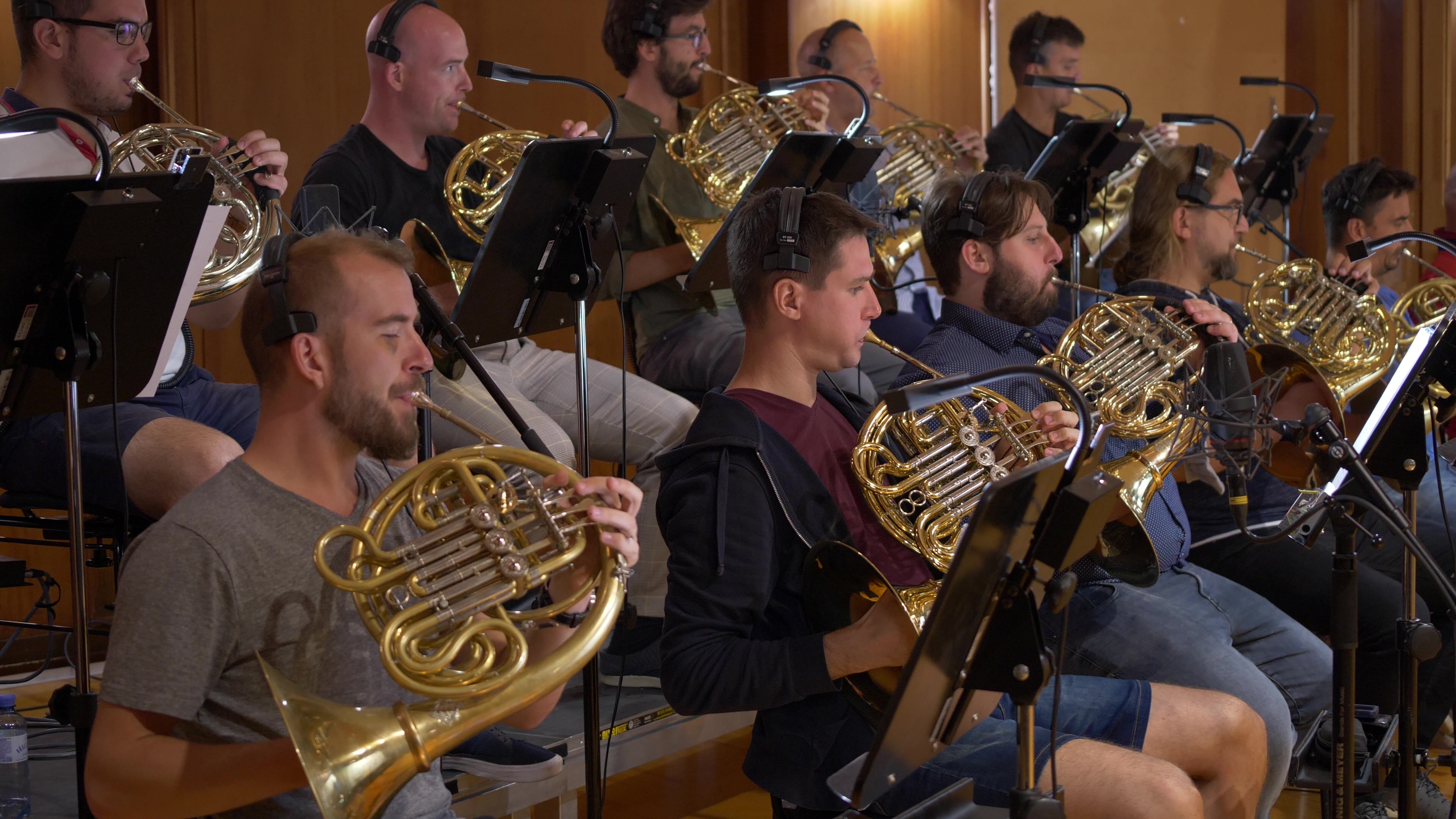 A group of male musicians wearing headphones playing French horns in an orchestra or recording studio, with music stands and microphones.