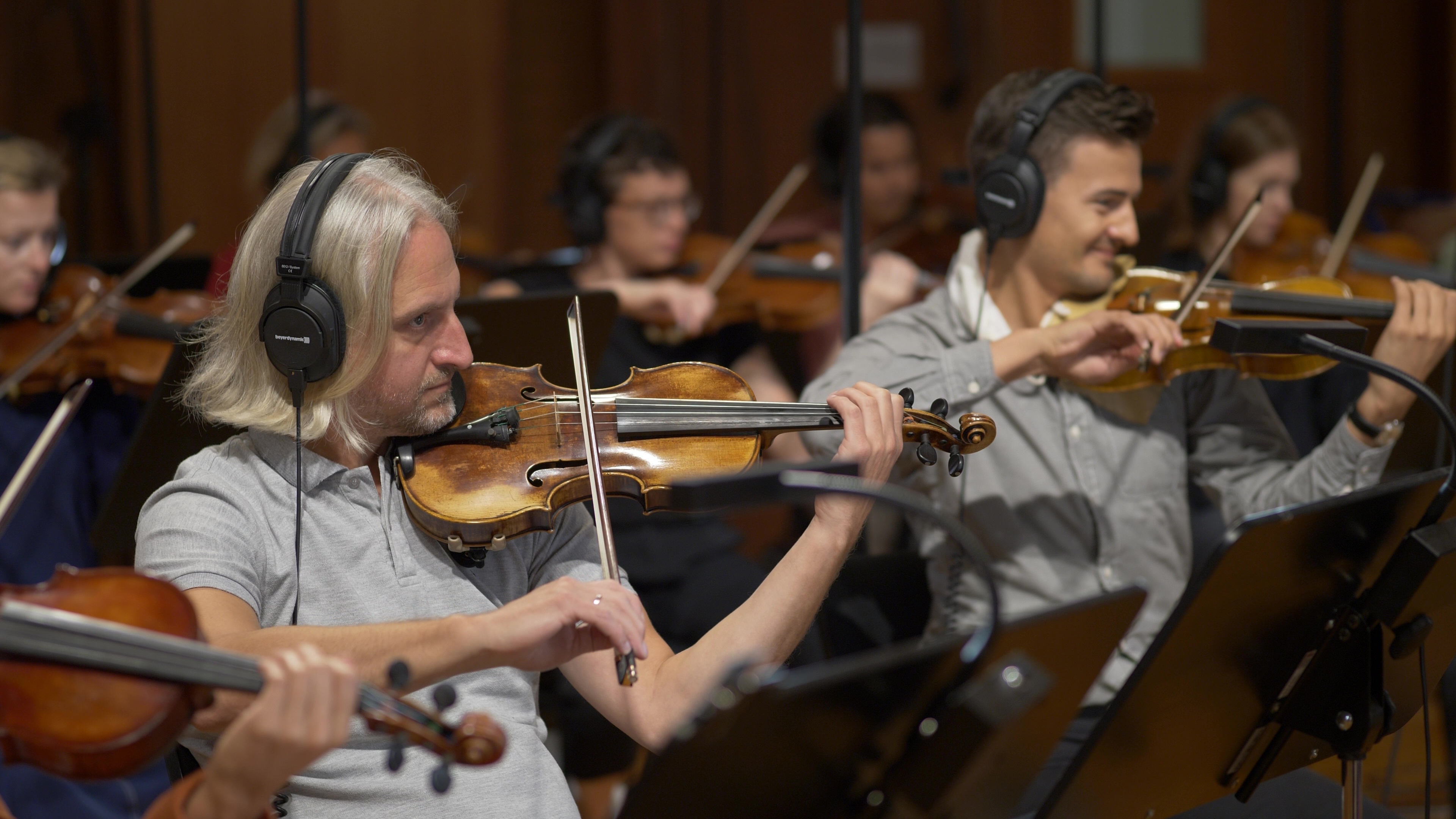 Musicians playing violins with headphones in a recording studio, featuring a blonde man in a grey polo shirt with a violin.