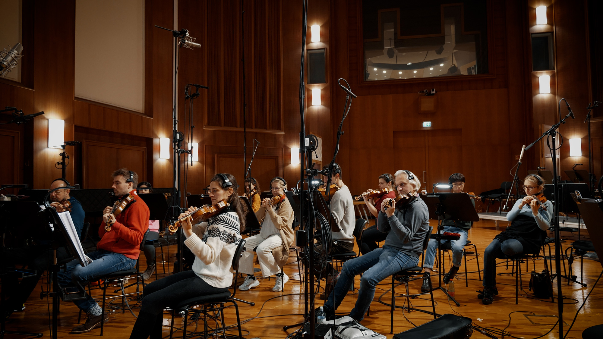 Orchestra musicians performing with violins and viola in a recording studio, surrounded by microphones and sheet music stands.