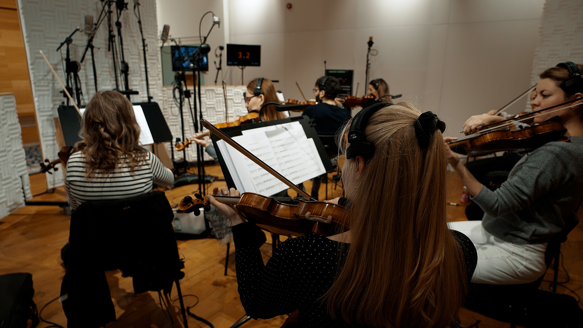 Orchestra members playing violins in a professional recording studio with microphones and sheet music stands.