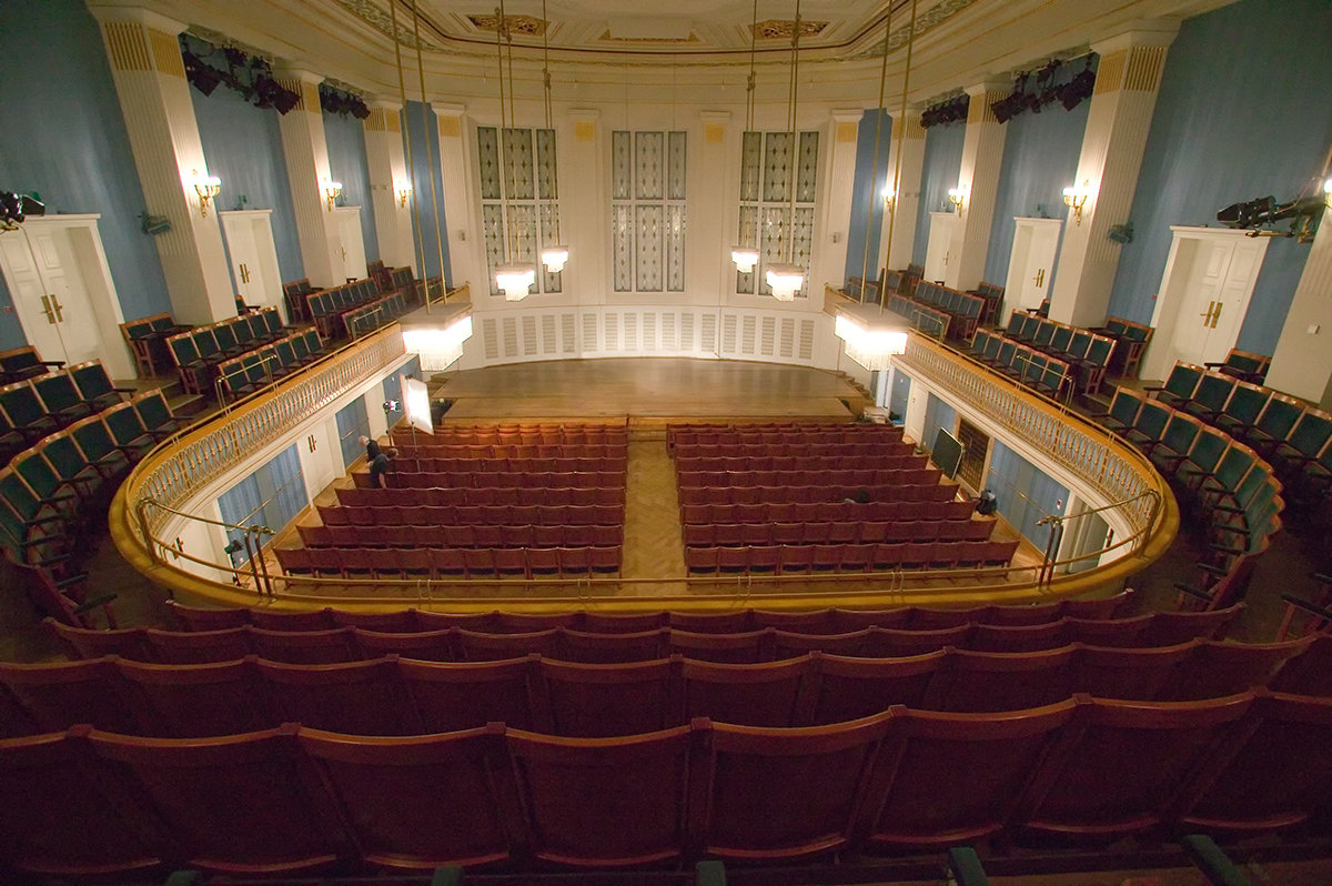 Large, empty concert hall or auditorium with a central stage, two sections of red seats on the ground floor, and two curved balconies with green seats. The walls are light blue and white with tall windows and sconces, and three large modern chandeliers hang over the stage.
