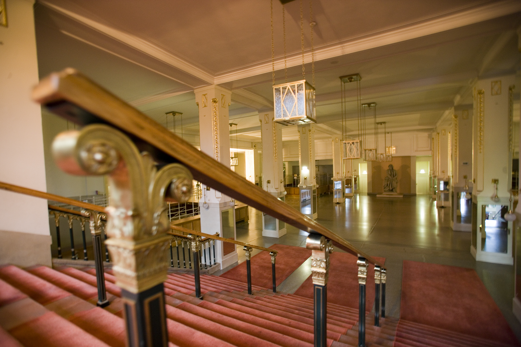 Grand ornate staircase with gold and black banisters, leading down to a brightly lit, expansive hall with tall white columns, gold accents, hanging cube-shaped lanterns, and a statue in the distance.