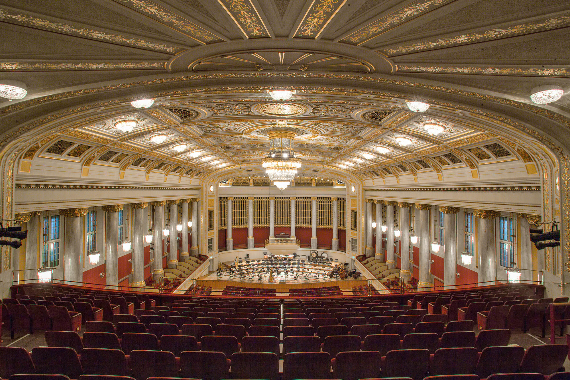 Grand concert hall interior with rows of empty red seats, a large orchestra stage, classical columns, and ornate gold and white architectural details.