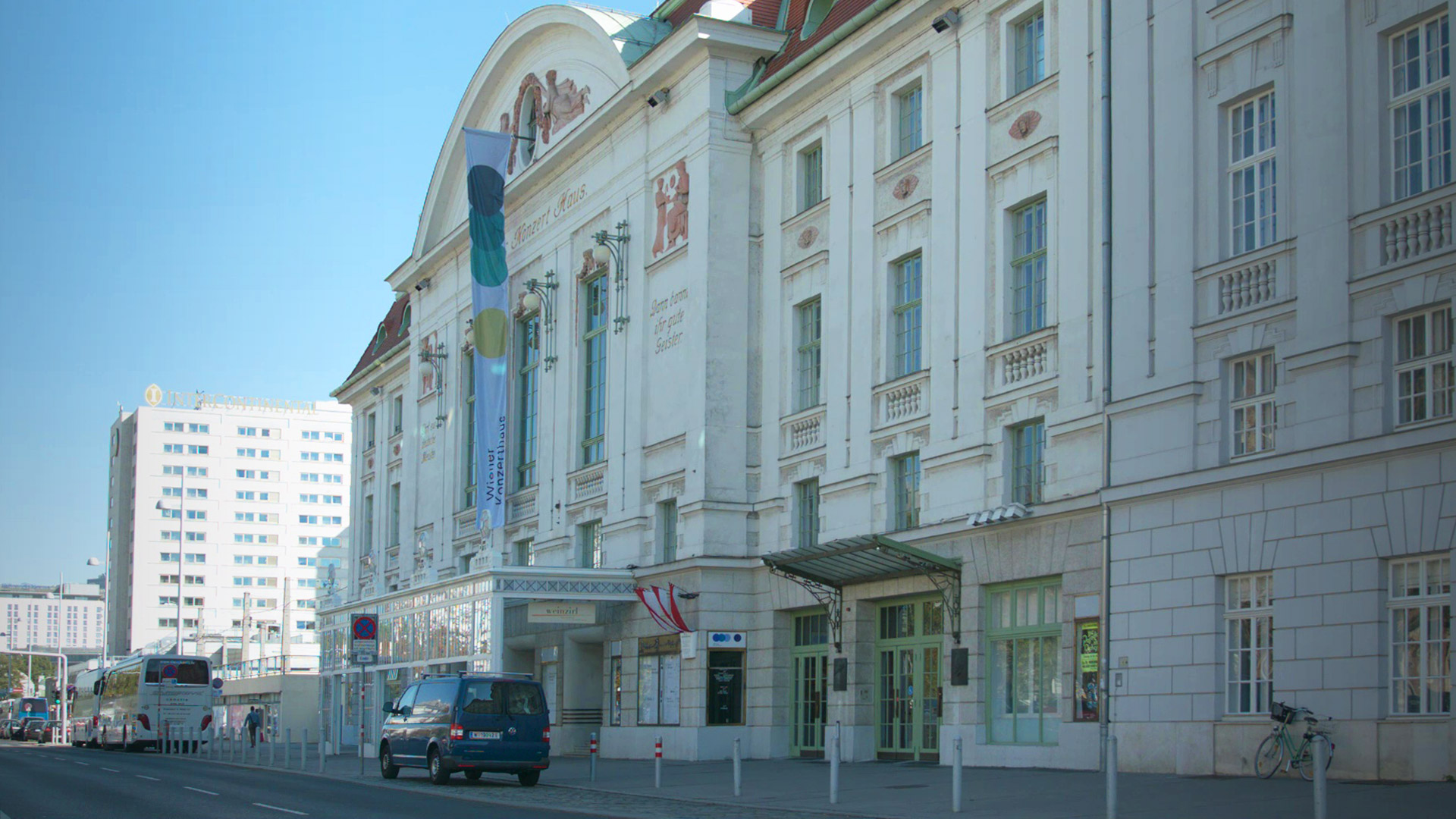 Wiener Konzerthaus in Vienna, Austria, with a colorful banner on its facade, street with vehicles, and a large white modern building in the background.
