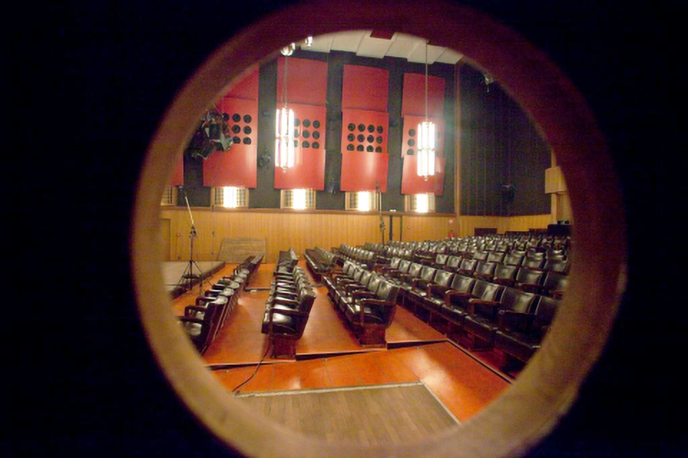 View through an opening of a large, empty auditorium with rows of brown seats, an orange floor, red wall panels, and illuminated hanging lamps.