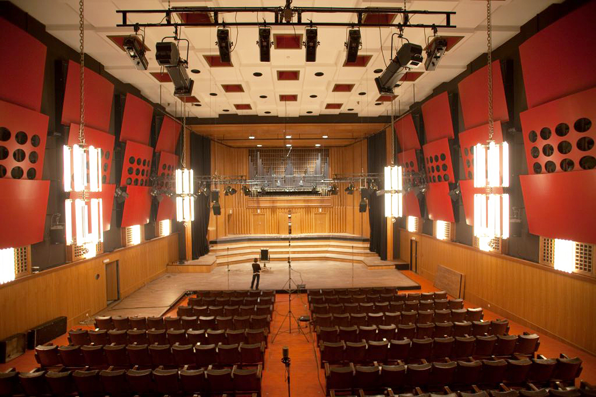 Wide view of a large concert hall or auditorium with rows of empty seats, a stage, and intricate acoustic wall panels.