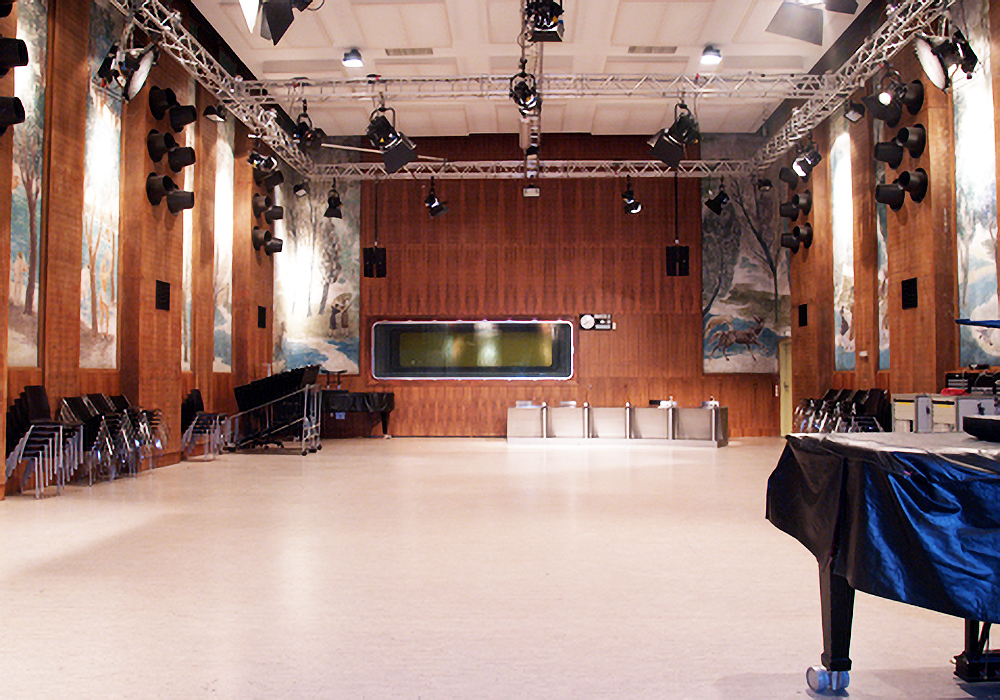 Concert hall interior with stage lighting, wooden paneling, nature murals, stacked chairs, piano, and sound mixing equipment.
