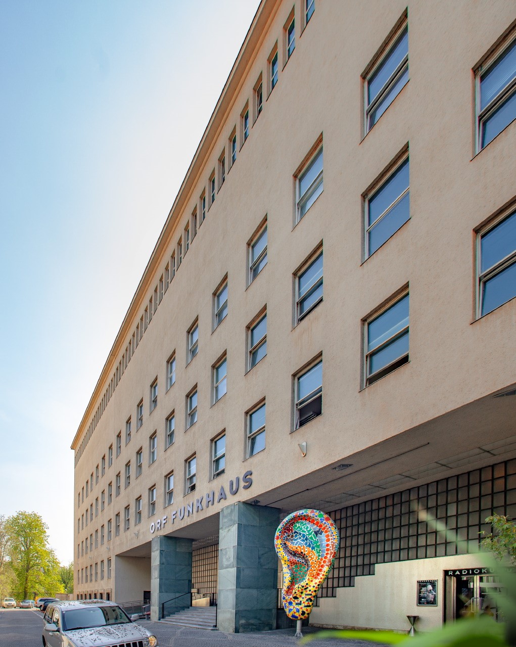 Exterior view of ORF Funkhaus building with ORF FUNKHAUS sign, multiple windows, and a colorful ear sculpture mosaic in the foreground