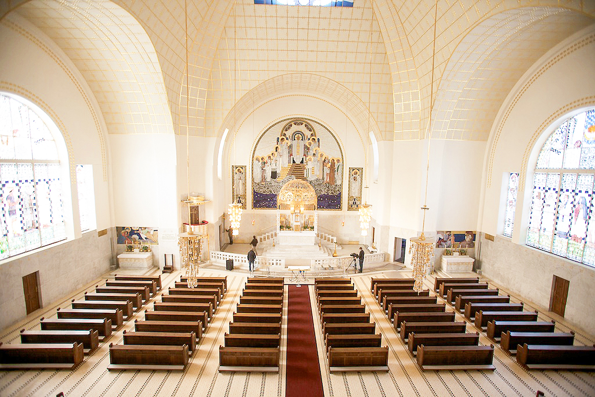 Ornate church interior with rows of wooden pews, high vaulted ceilings, stained glass windows, and a mosaic altar.