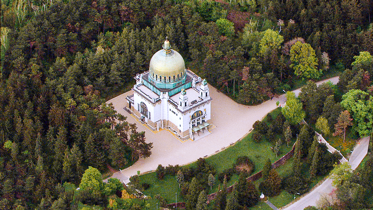 Aerial view of Steinhof Church in Vienna, Austria, showcasing its white exterior and large golden dome surrounded by dense green forest trees and walking paths.