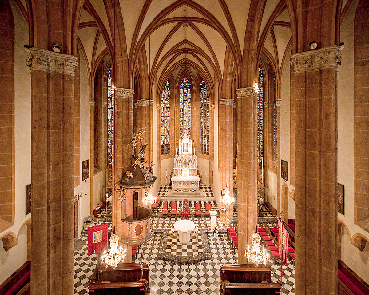 Interior view of a grand Gothic cathedral or church showing the altar, stained glass windows, large stone columns, archways, and checkered tile flooring.