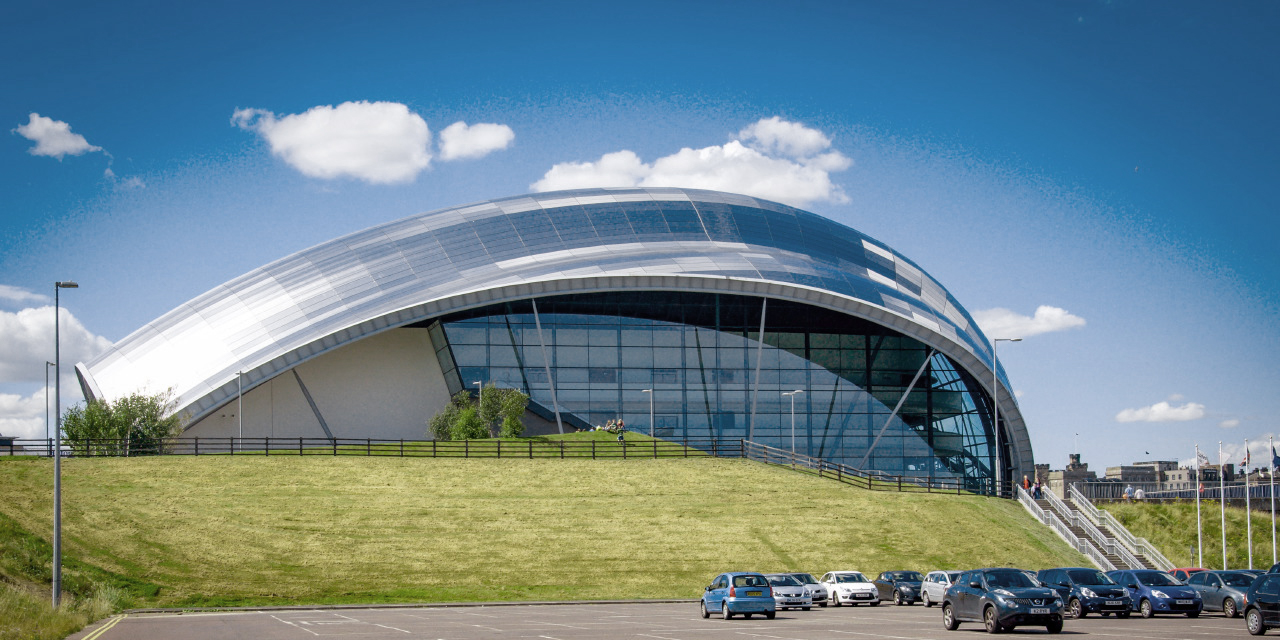 Sage Gateshead concert venue, a modern curved glass and metal building, with a green grassy hill leading up to it from a parking lot filled with cars under a bright blue sky with white clouds.