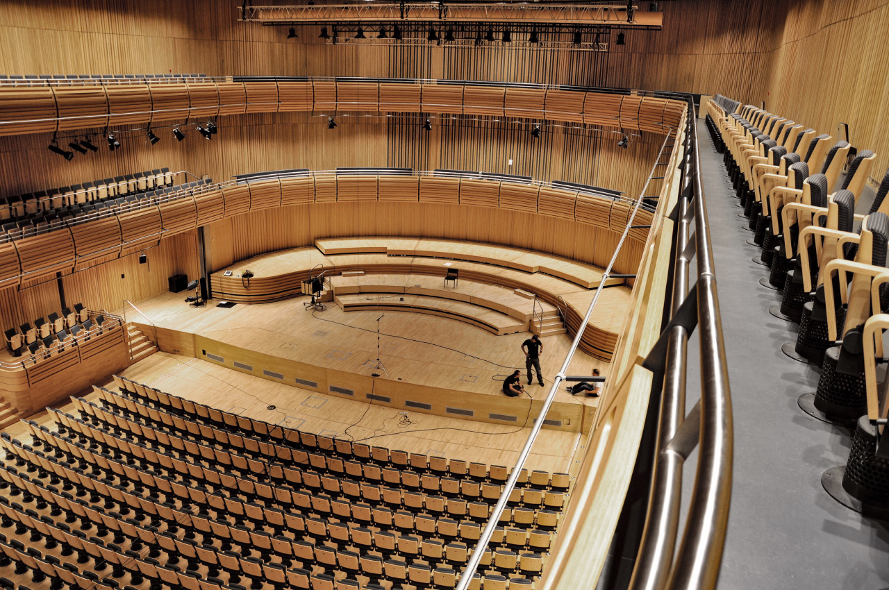 Spacious modern concert hall or auditorium with light wood paneling, rows of empty seats, a large stage with three people, and overhead lighting.