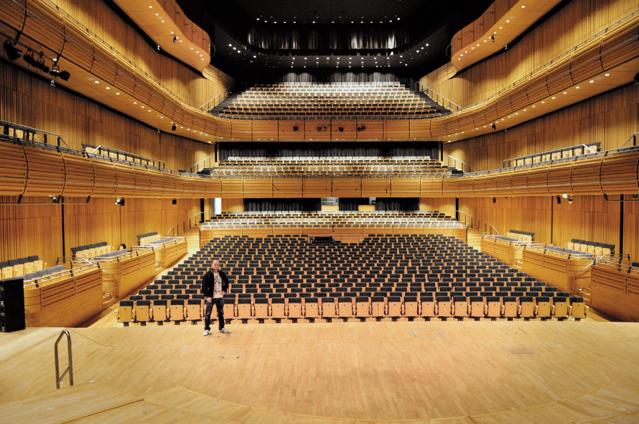 Wide view of an empty modern concert hall or auditorium with tiered seating, featuring light wood paneling, black seats, and a large stage. A man stands on the stage at the center.