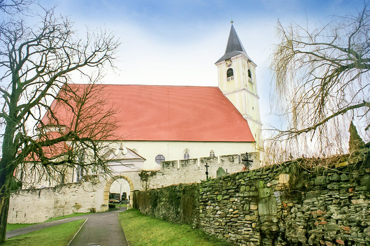 Historic church with red roof and tall bell tower behind a stone wall and bare trees, a pathway leads to a gated entrance.