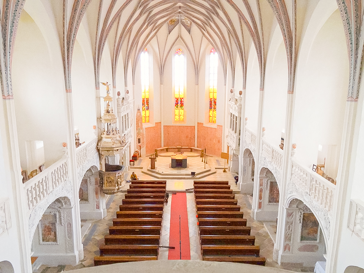 Interior view from above of a bright church nave with arched ceilings, stained-glass windows, wooden pews, an ornate pulpit, and a central altar.