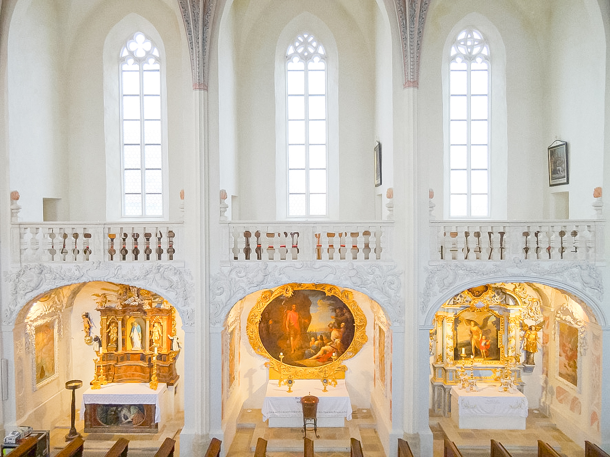 Interior view of a European church nave, featuring three alcoves with altars and religious artwork below a white balcony with three tall arched windows.