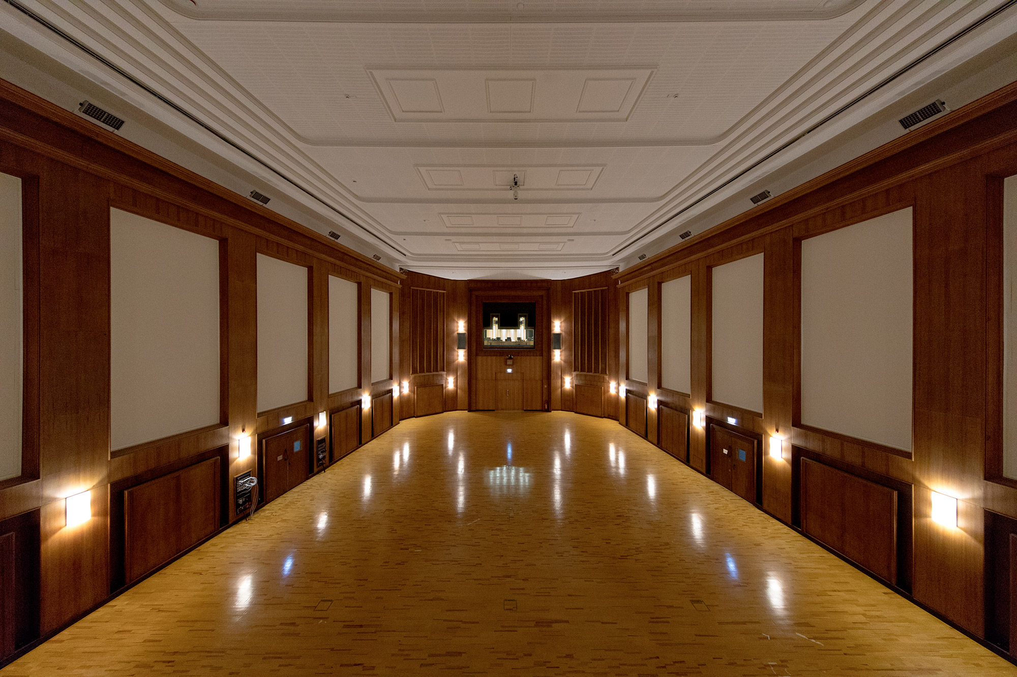 Conference room with polished wooden floors reflecting warm wall lights, cream wall panels, and a distinctive curved far wall featuring a podium and screen.