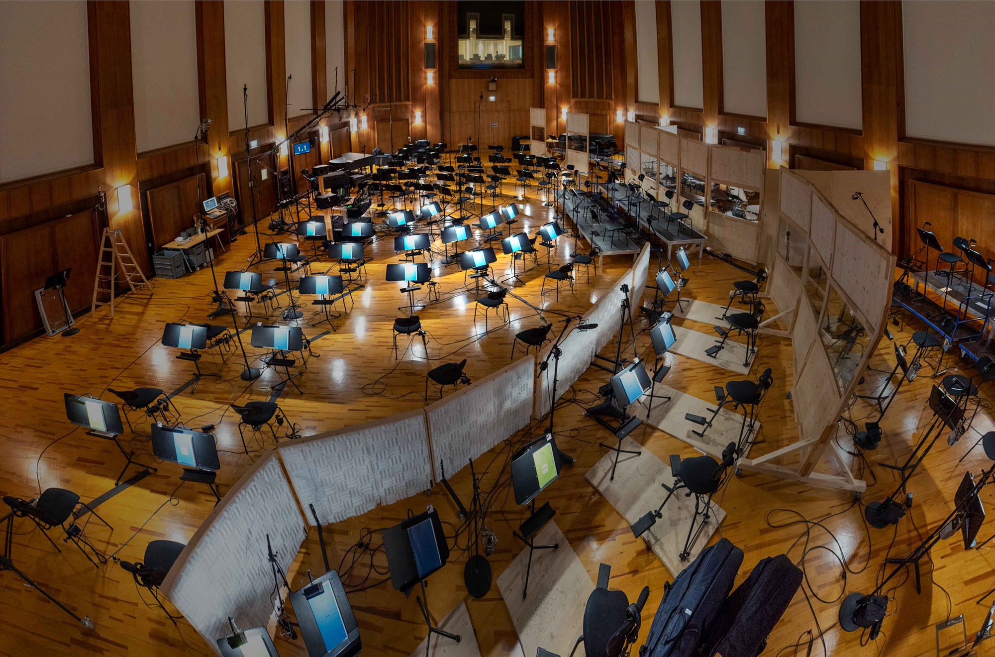 Large orchestral recording studio with chairs, music stands with screens, microphones, and sound baffles on a wooden floor, ready for musicians.