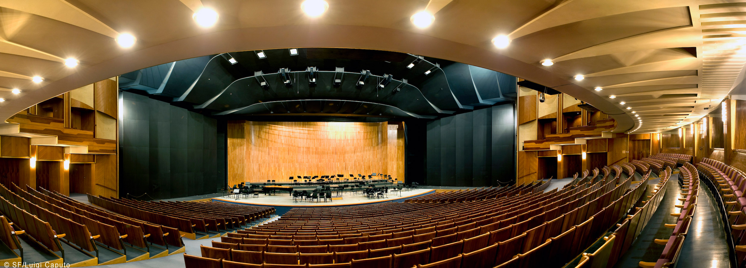 Panoramic view of a modern theater auditorium with rows of empty wooden seats, a stage set with music stands and instruments, and sophisticated acoustic paneling and lighting.
