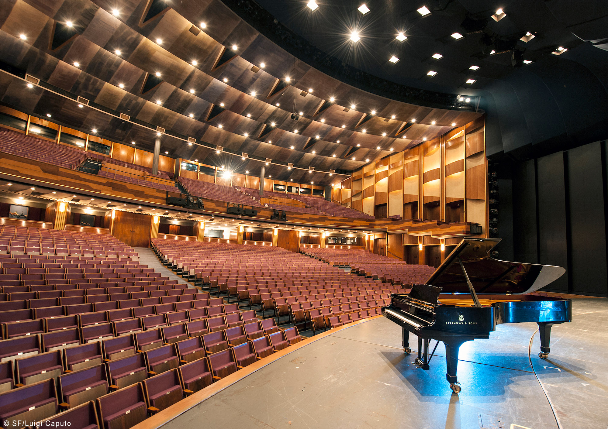 Wide view of a large, empty modern concert hall or auditorium with rows of burgundy seats, a stage with a black Steinway & Sons grand piano, and illuminated acoustic ceiling panels.