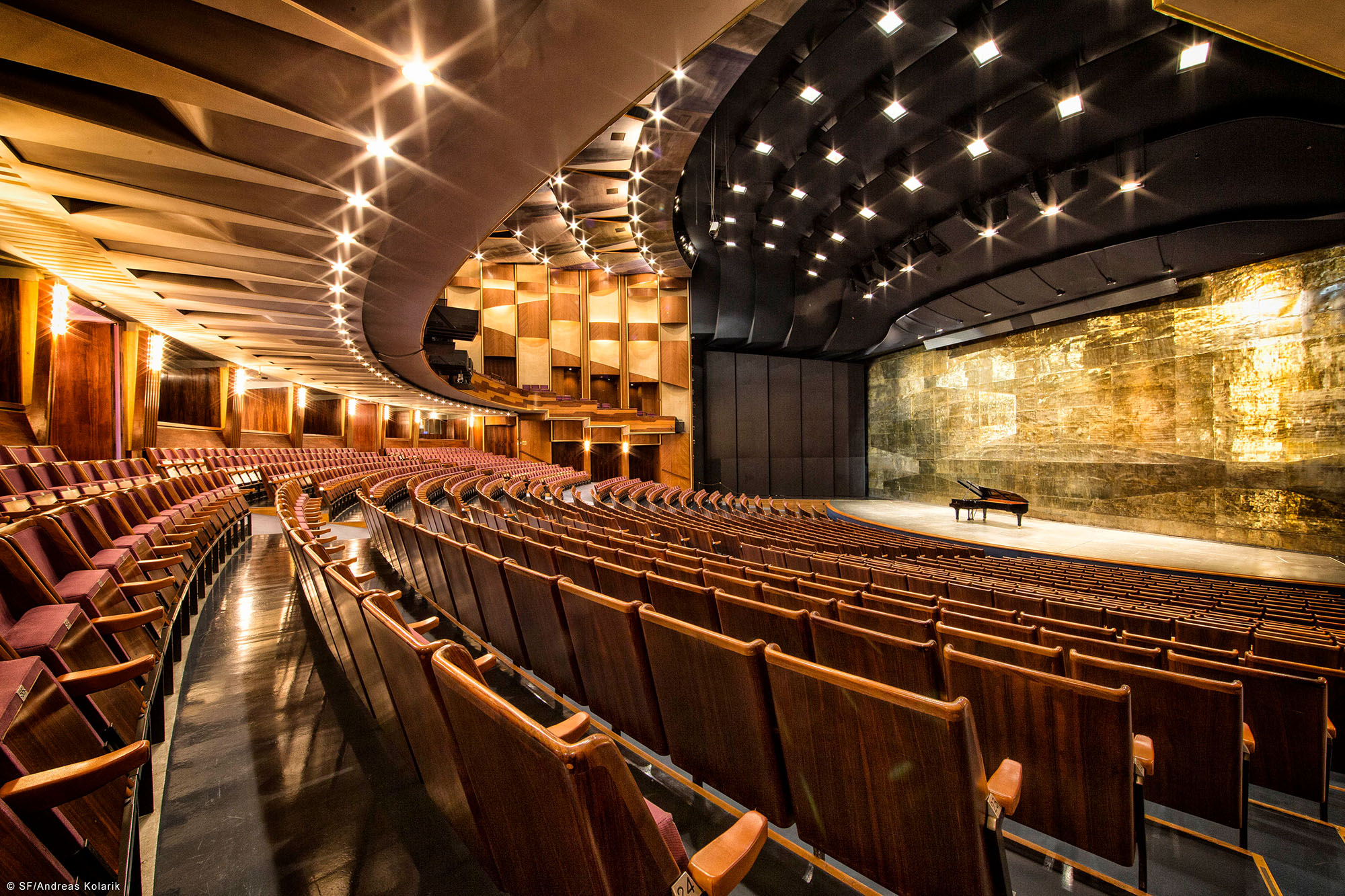 Spacious opera house auditorium with rows of elegant red seats, ornate wooden walls, bright overhead lights, and a grand piano centered on the stage with a textured golden backdrop.