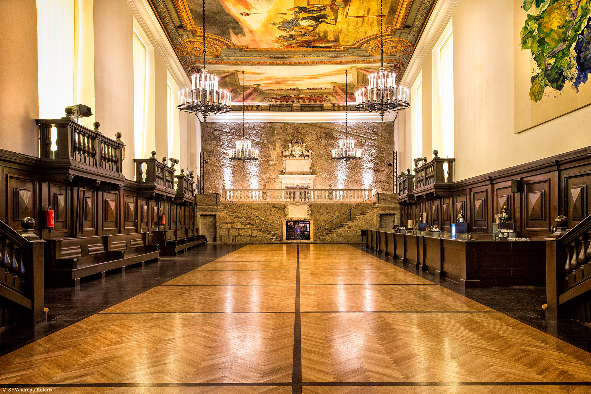Grand hall with ornate wooden paneling, a large parquet floor, two-tiered stone staircase leading to a decorative archway, illuminated by multiple chandeliers and natural light from tall windows, featuring a painted ceiling mural.