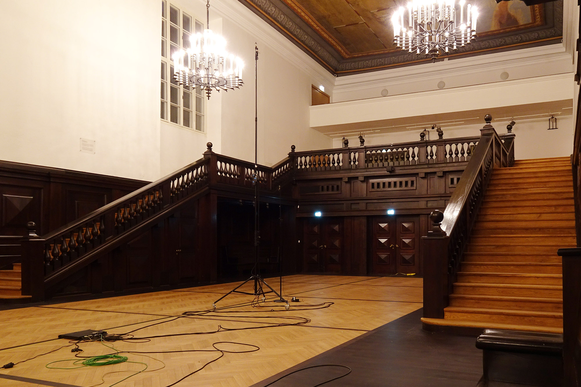 Grand hall with parquet floor, dark wood paneled walls, two prominent wooden staircases, three large chandeliers, and a high ceiling.