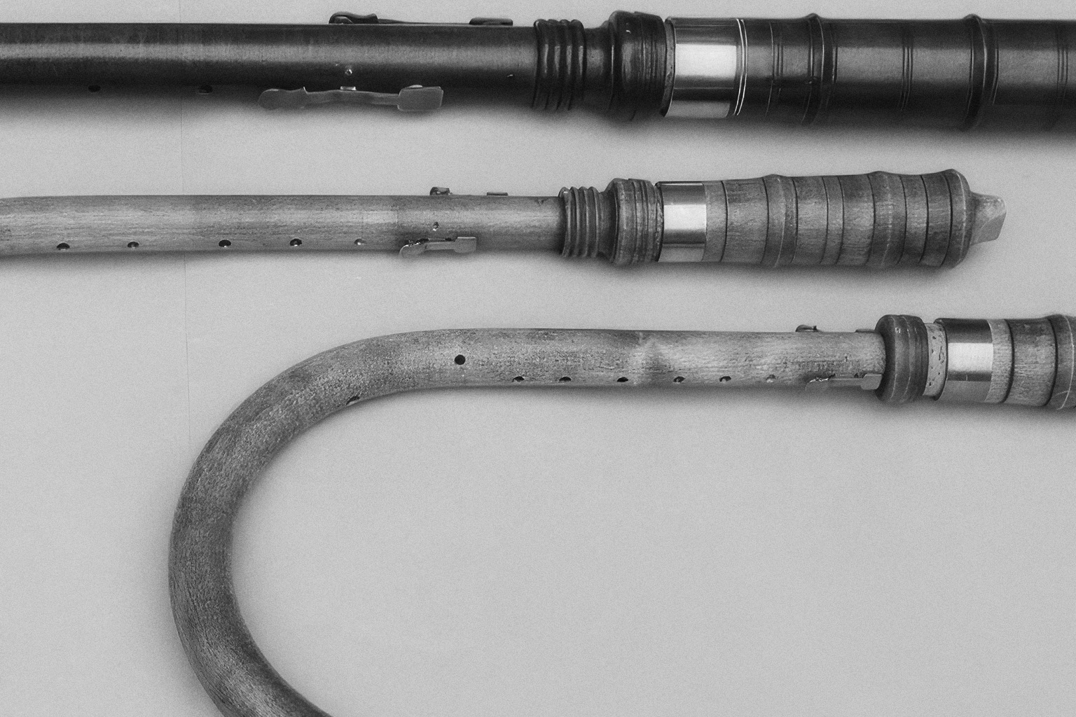 Black and white image showing three bagpipe chanters laid horizontally. The top chanter is dark and appears to be made of ebony. The middle chanter is lighter, made of wood, and has a ribbed section. The bottom chanter is also wooden, curved at one end, and features several holes along its length. All chanters have visible metal bands and keys.