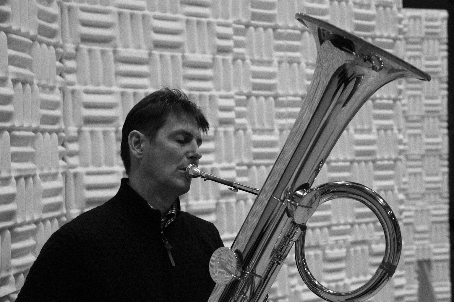 Black and white image of a man playing a shiny tuba in an acoustically treated room, focusing on his engaged expression and the instrument's reflective surface.
