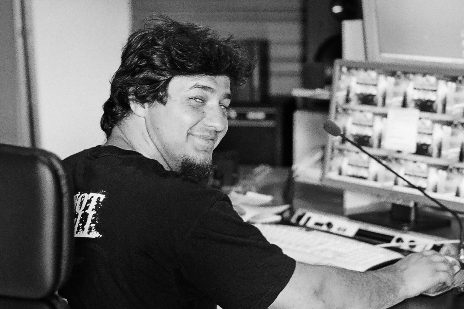 Black and white photo of a smiling man with curly hair and a goatee, sitting at a desk with a computer monitor, microphone, and keyboard, looking over his shoulder toward the viewer.