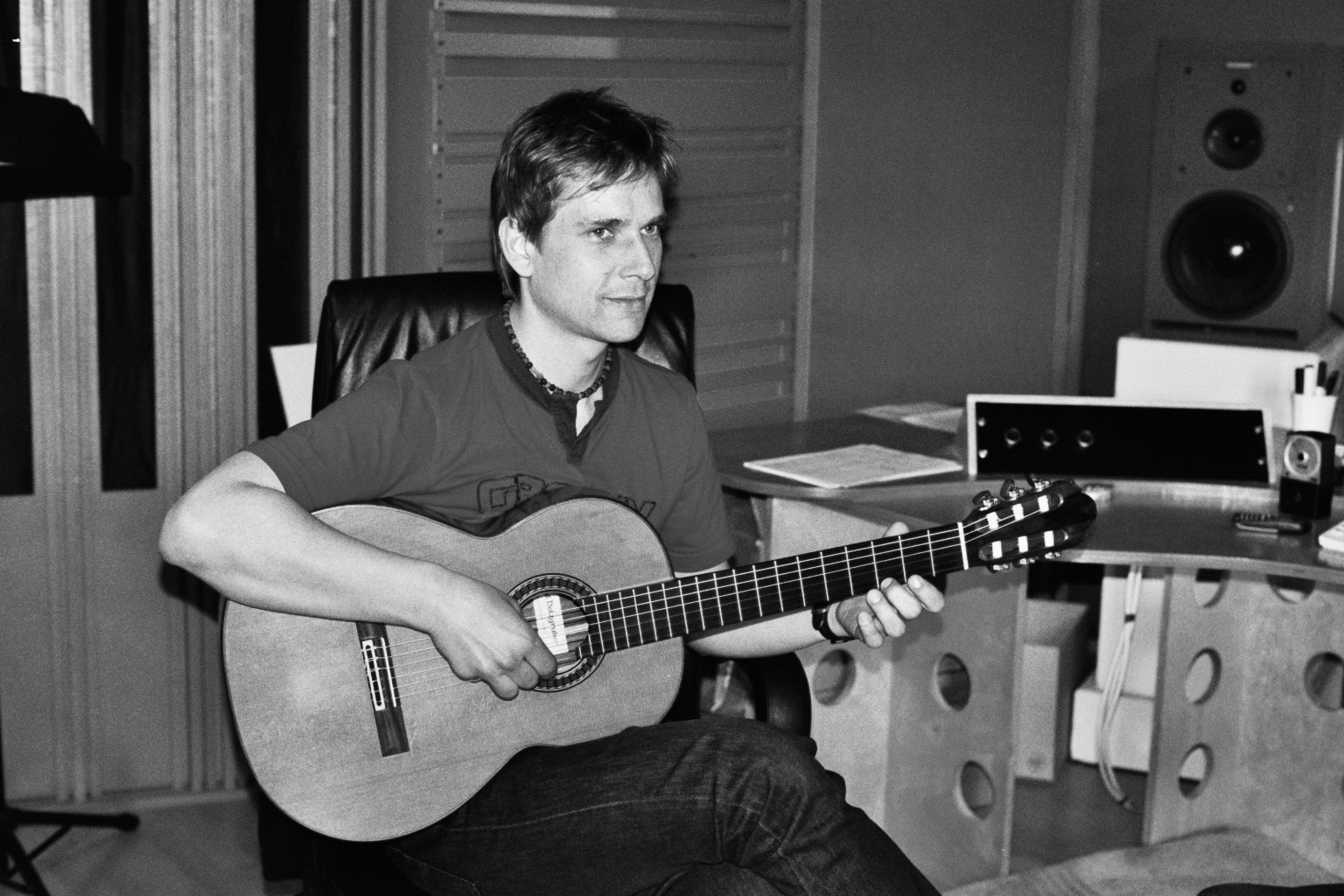Black and white image of a man playing an acoustic guitar indoors, likely in a recording studio. He is seated, looking to the right with a focused expression. Studio equipment is visible in the background.