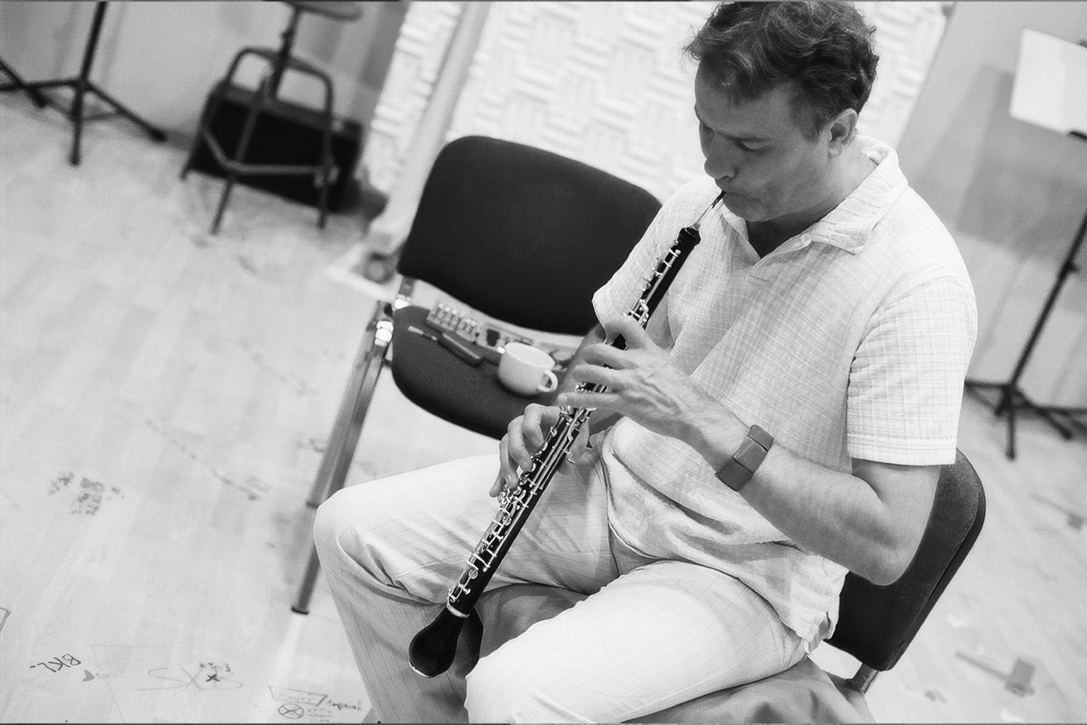 Black and white medium close-up of a man seated, playing an oboe with focused intensity, his hands positioned on the intricate keys of the instrument. The surrounding studio environment suggests a musical performance or practice session.