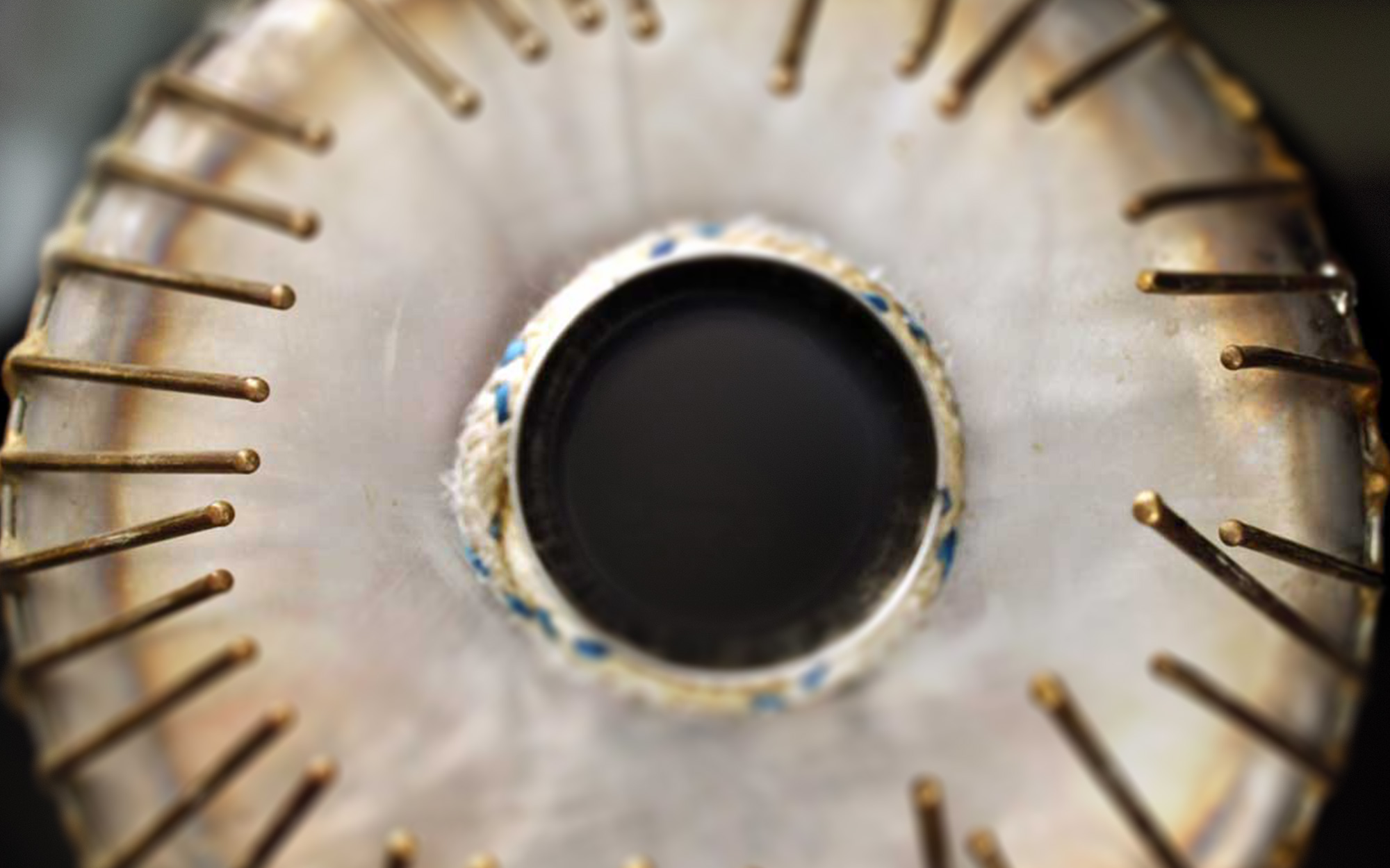 Close-up of a circular metal object with radiating brass rods resembling a traditional Indian musical instrument drum, possibly a tabla or mridangam, featuring a central dark circular opening wrapped in rope.