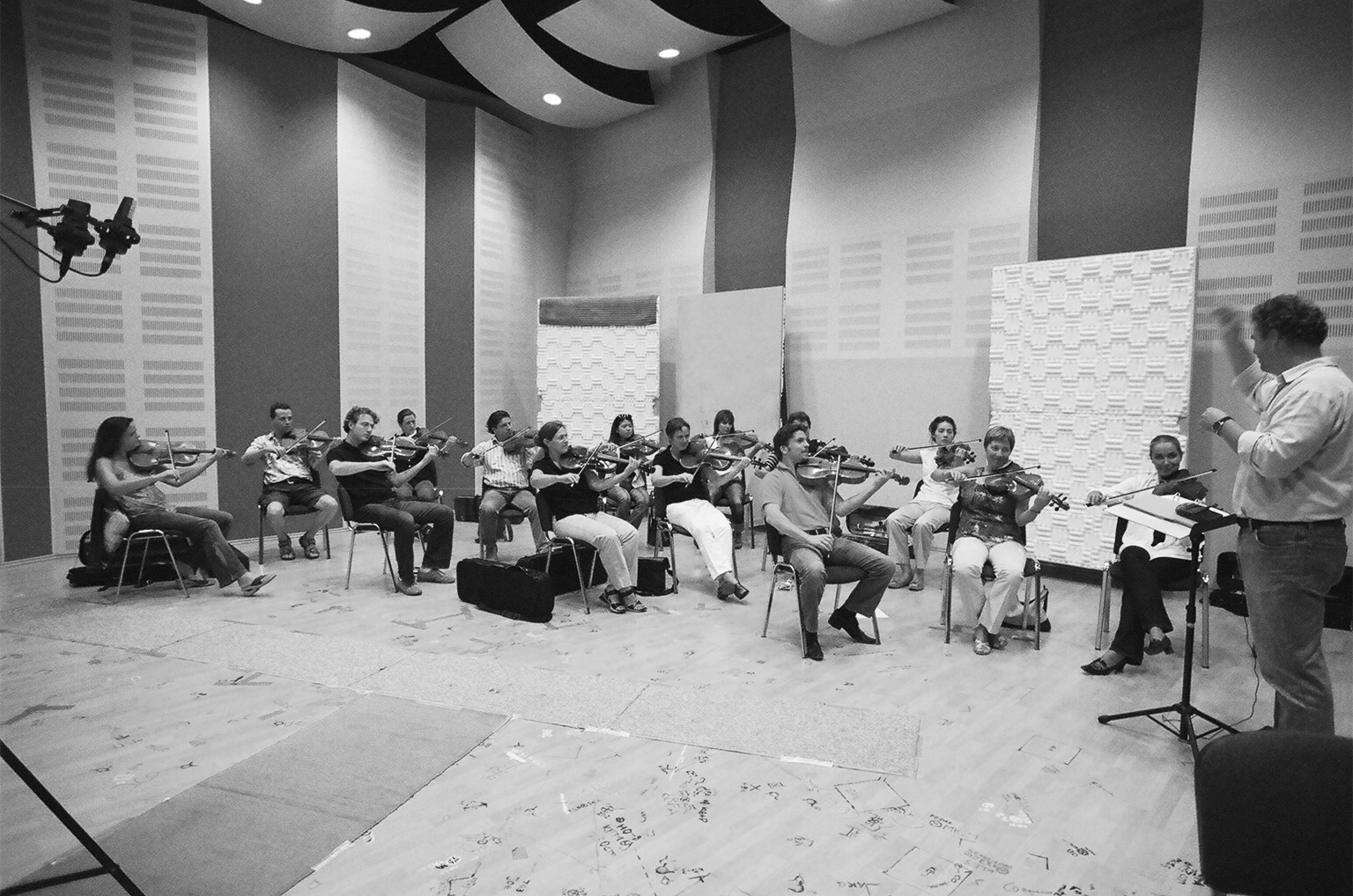 Black and white image of a string orchestra playing violins in a recording studio, with a conductor leading them.