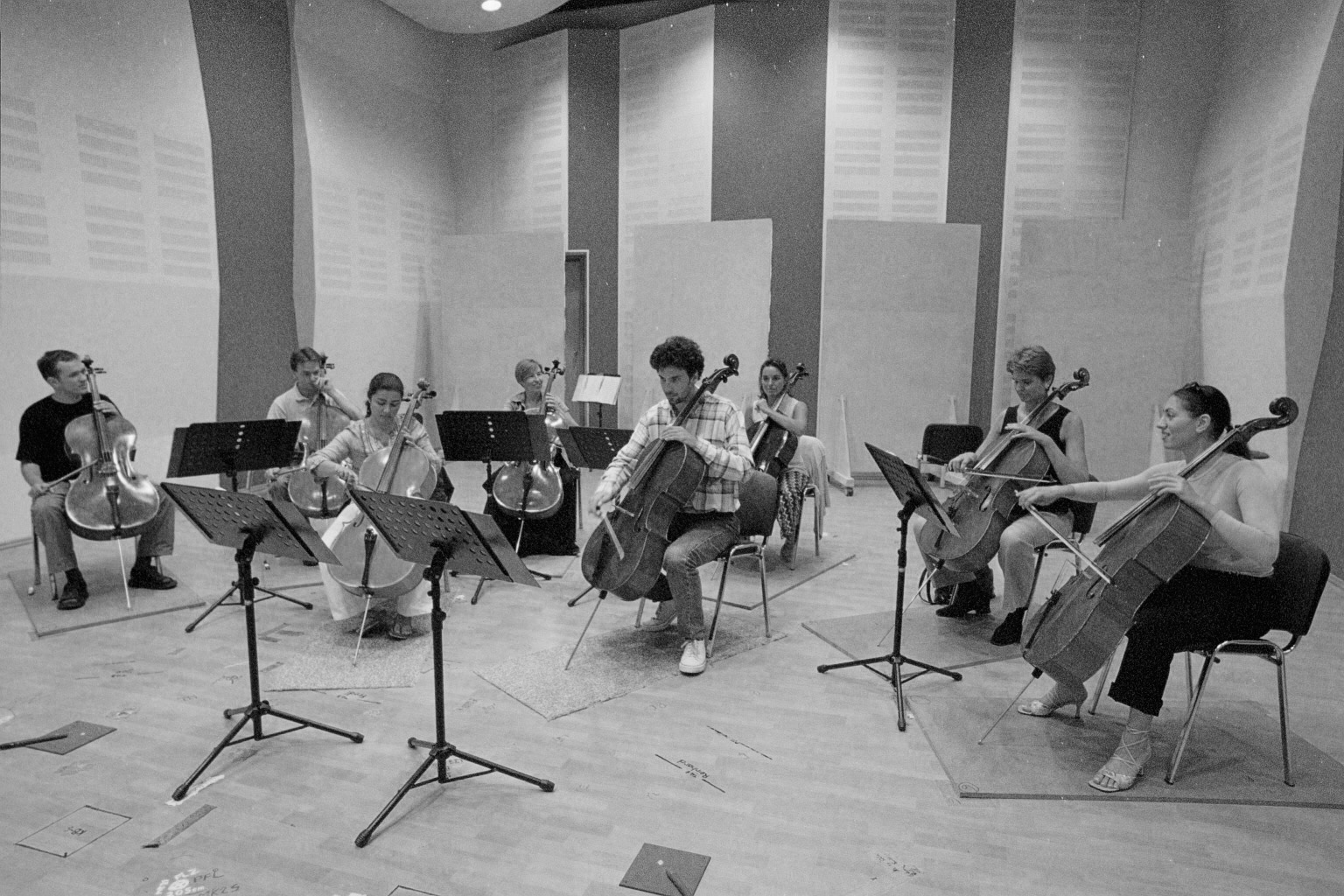 Black and white photo of a group of six musicians practicing cello in a rehearsal room.