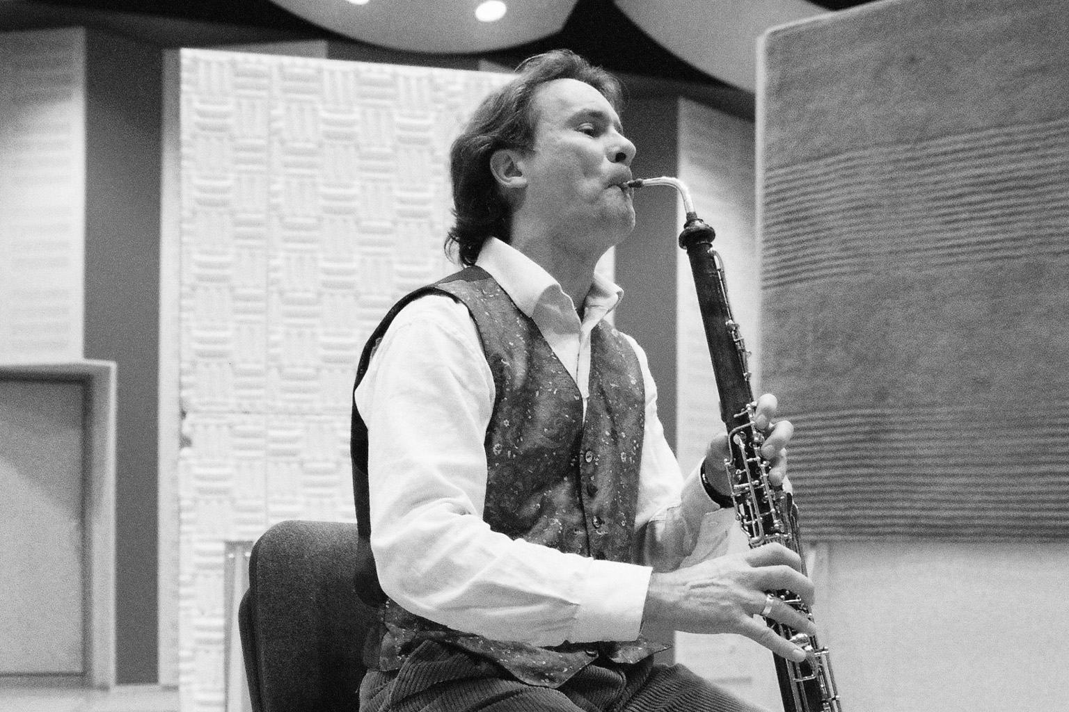 Black and white angled shot of a male musician playing an oboe with intensity, wearing a white shirt and patterned vest, in a studio with sound-dampening panels.