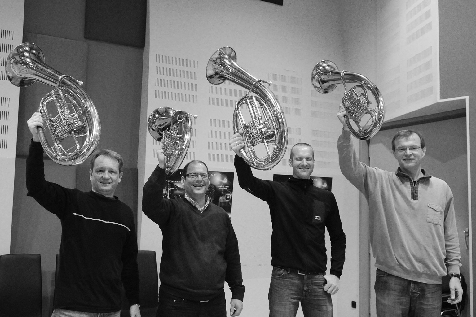 Black and white image of four men smiling and holding up shiny brass coiled musical instruments resembling small tubas or euphoniums.