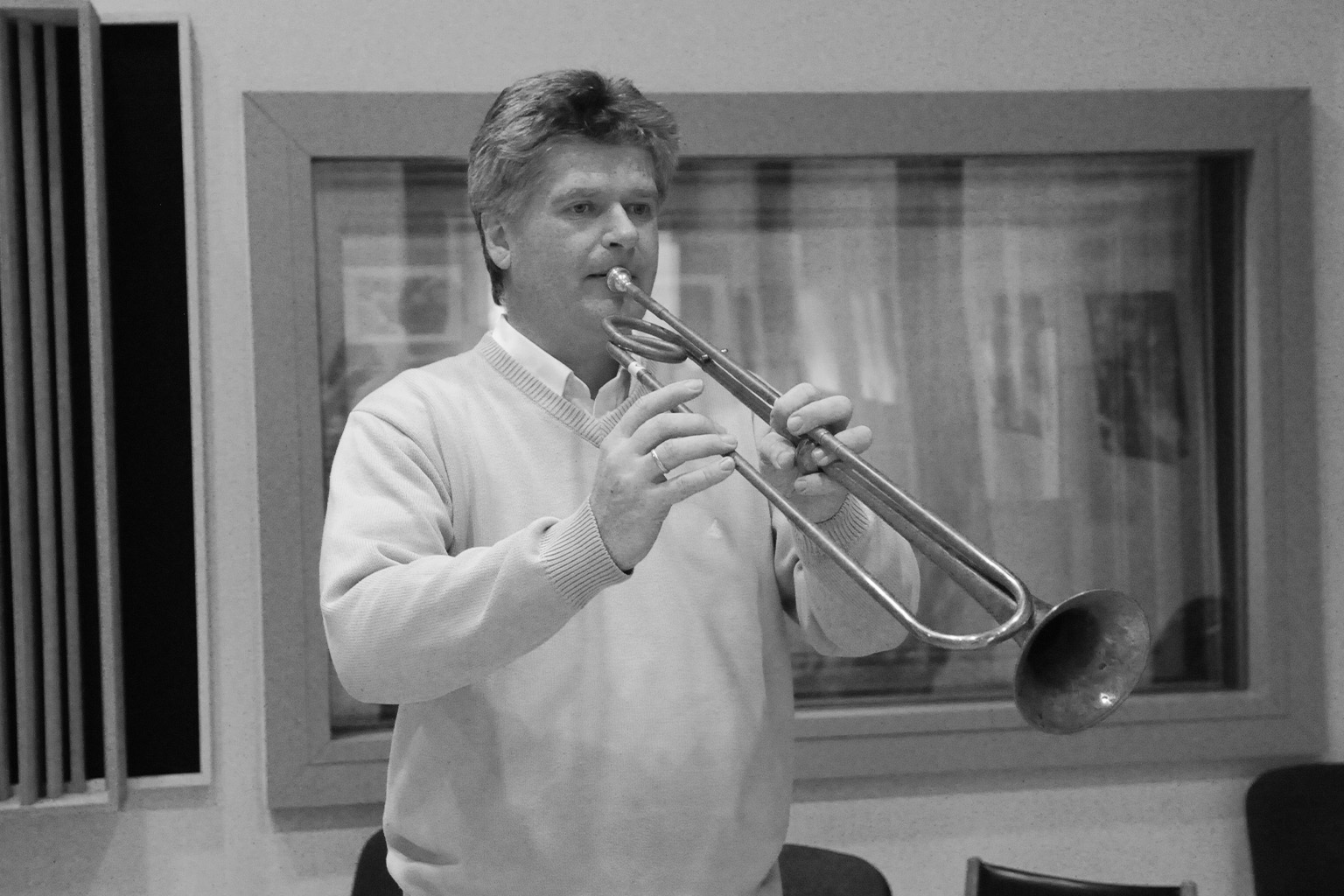 Black and white capture of a man playing the trombone indoors, wearing a light-colored V-neck sweater over a collared shirt.