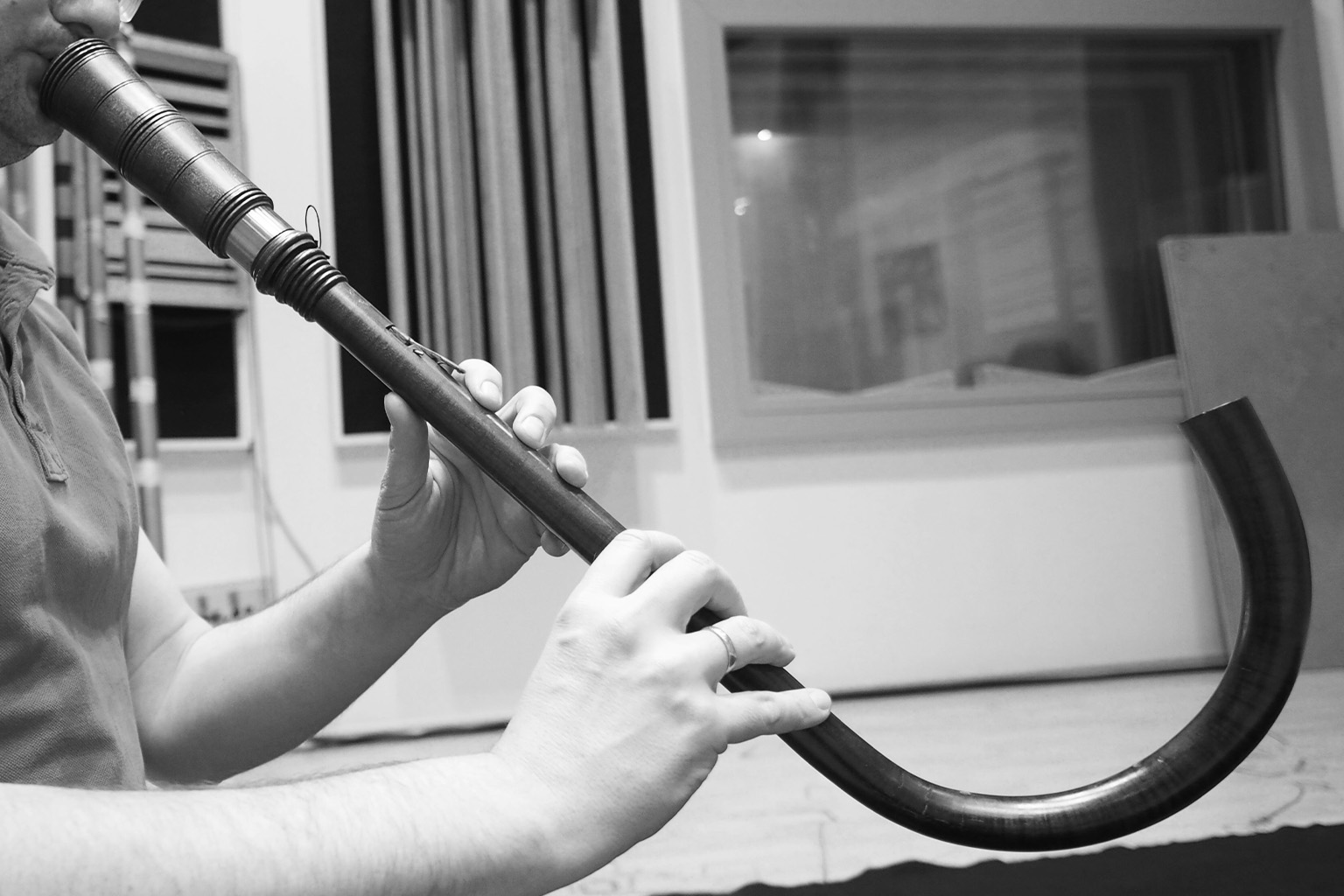 Black and white image of a person playing a traditional curved wind instrument, possibly a cornett or medieval horn, with focused hands and mouth.